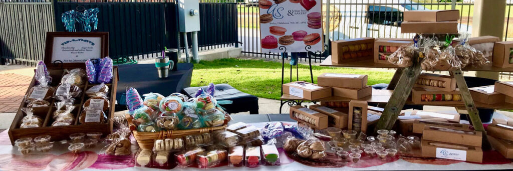 Artisan Sweets Bakery booth at the Rose District Farmers Market in Broken Arrow Oklahoma — macaron boxes, handmade cookies, and novelty treats on display.